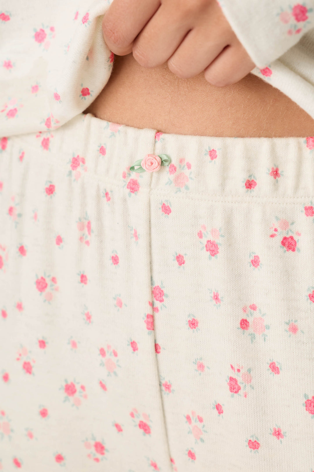 A close-up shows a person’s hand adjusting the waistband of the P.J. Salvage Romantic Rose Ditsy Rose Print Pajama Pants, featuring a cream background with pink floral patterns and a small pink button in the center.