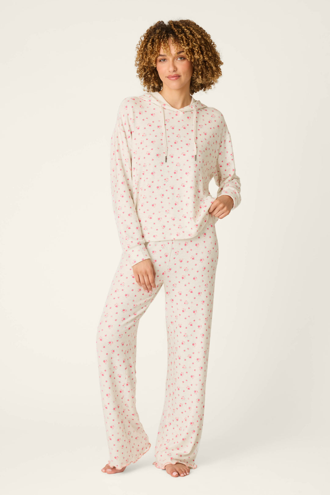 A person stands against a plain light background wearing the P.J. Salvage Romantic Rose Ditsy Rose Print Hoodie & Pants PJ Set, which features a hooded long-sleeve top and wide-leg pants in white with a small pink floral design.