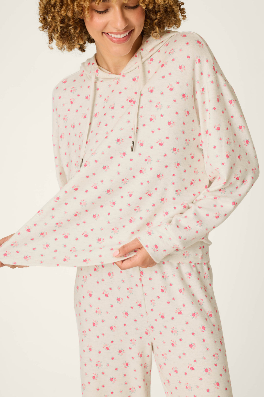 A person wearing the P.J. Salvage Romantic Rose Ditsy Rose Print Hoodie & Pants PJ Set, smiling and holding out the side of the cream-colored floral hoodie.