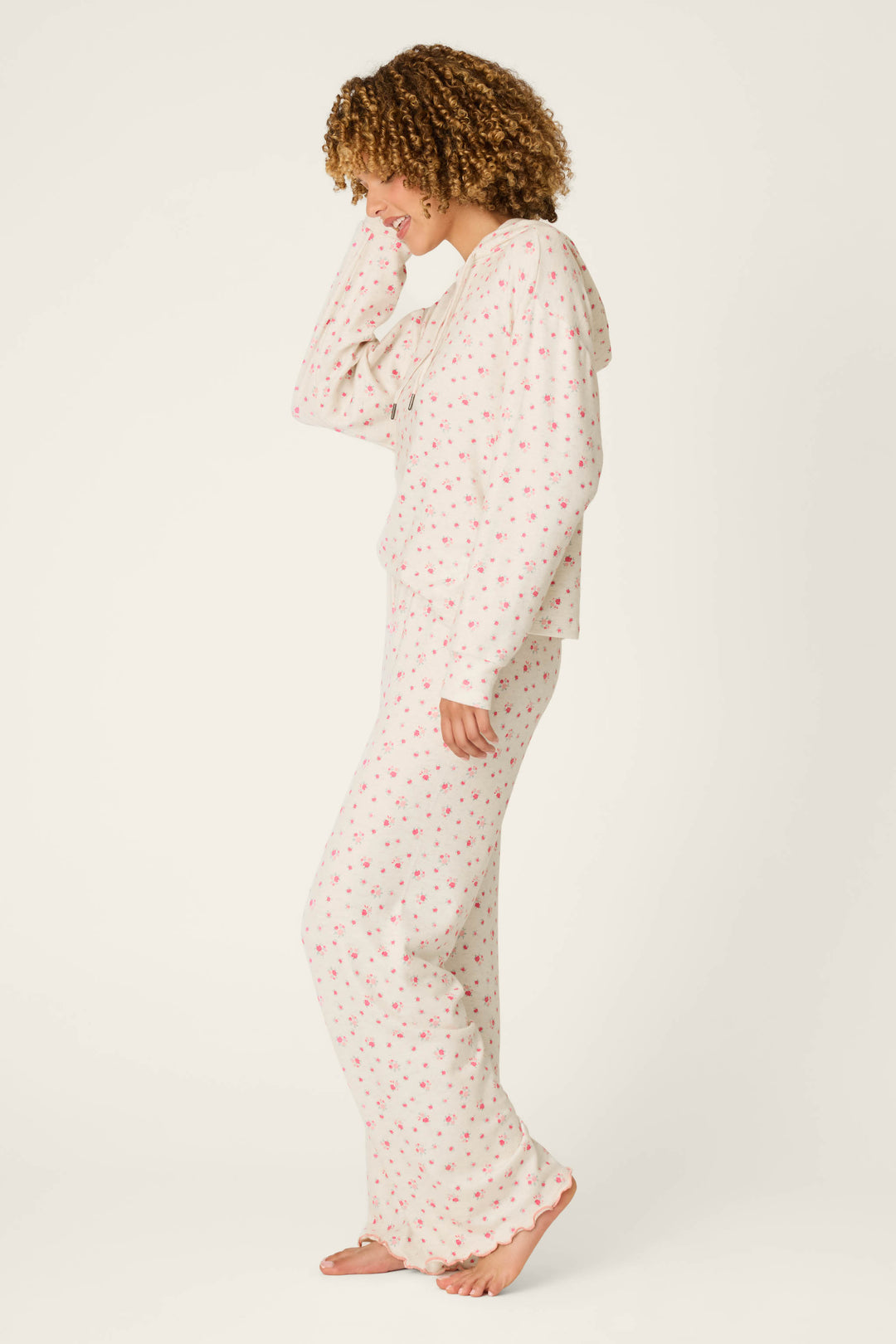 A woman wearing the P.J. Salvage Romantic Rose Ditsy Rose Print Hoodie & Pants PJ Set stands barefoot on a white background, smiling and looking down with one hand touching her hair.