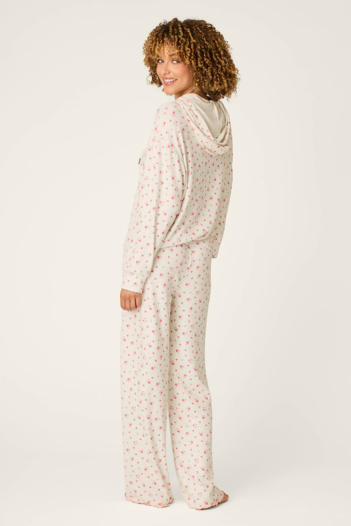 A woman with curly hair is wearing the P.J. Salvage Romantic Rose Ditsy Rose Print Hoodie & Pants PJ Set, featuring a white background with small pink and green floral prints. She stands smiling and is turned slightly to show the back of the outfit.
