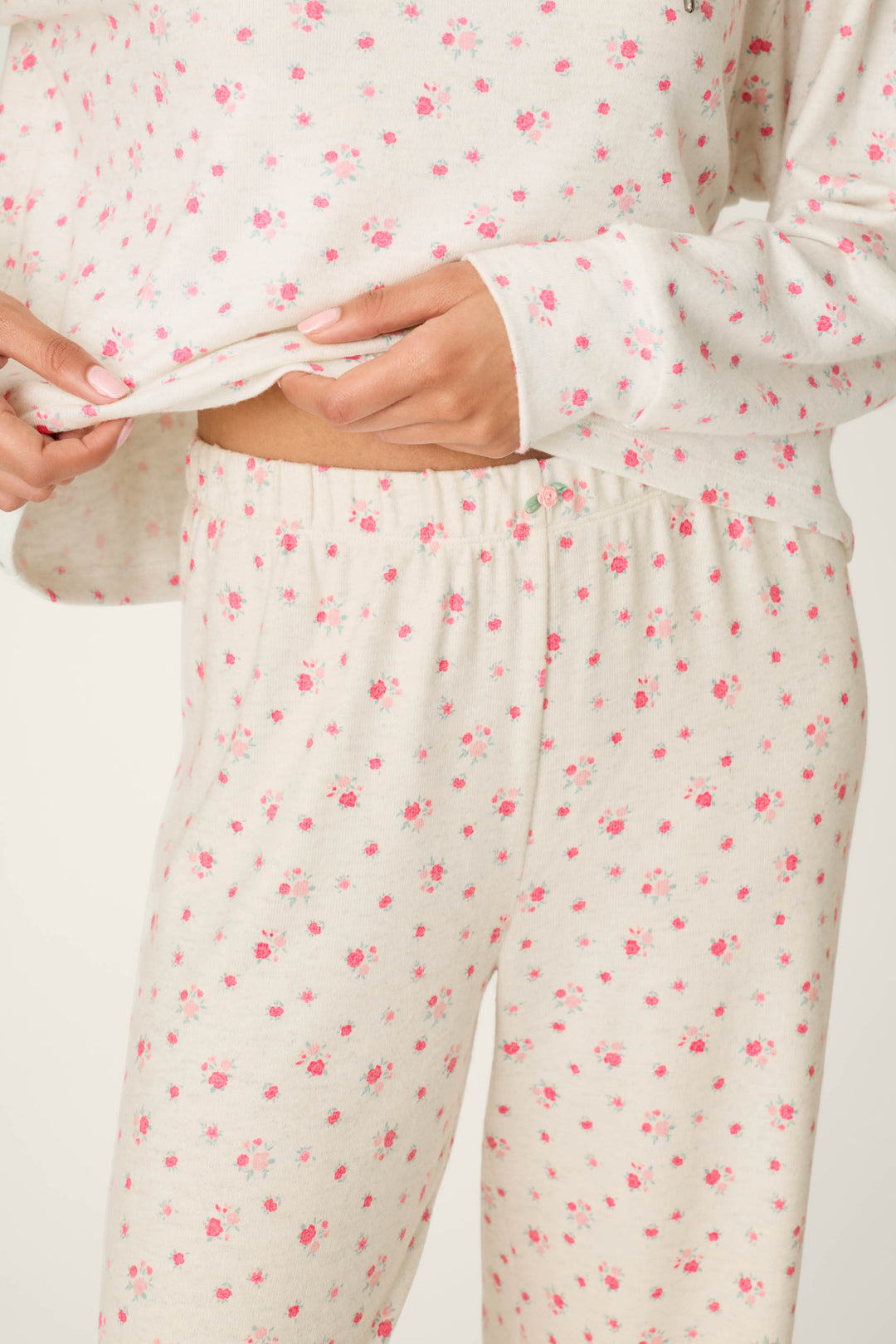 A person wearing the P.J. Salvage Romantic Rose Ditsy Rose Print Hoodie & Pants PJ Set holds the edge of their top, revealing the matching pants. The image focuses on their torso and hands.