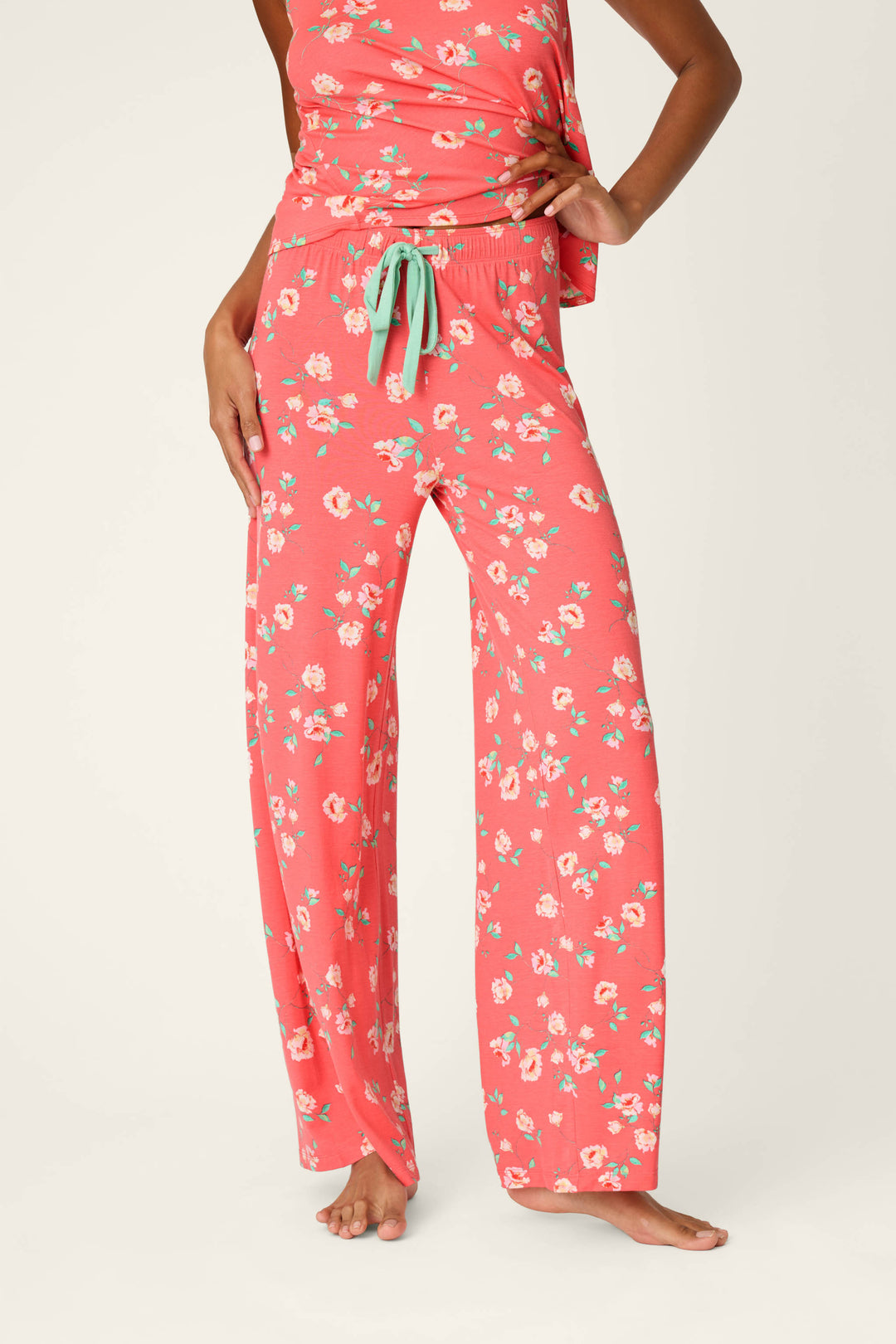A woman, barefoot, models P.J. Salvage Siesta Dreams Dark Coral Floral Print Pajama Pants with a green drawstring, made from soft modal jersey; only her lower body and part of her torso are visible against a plain background.