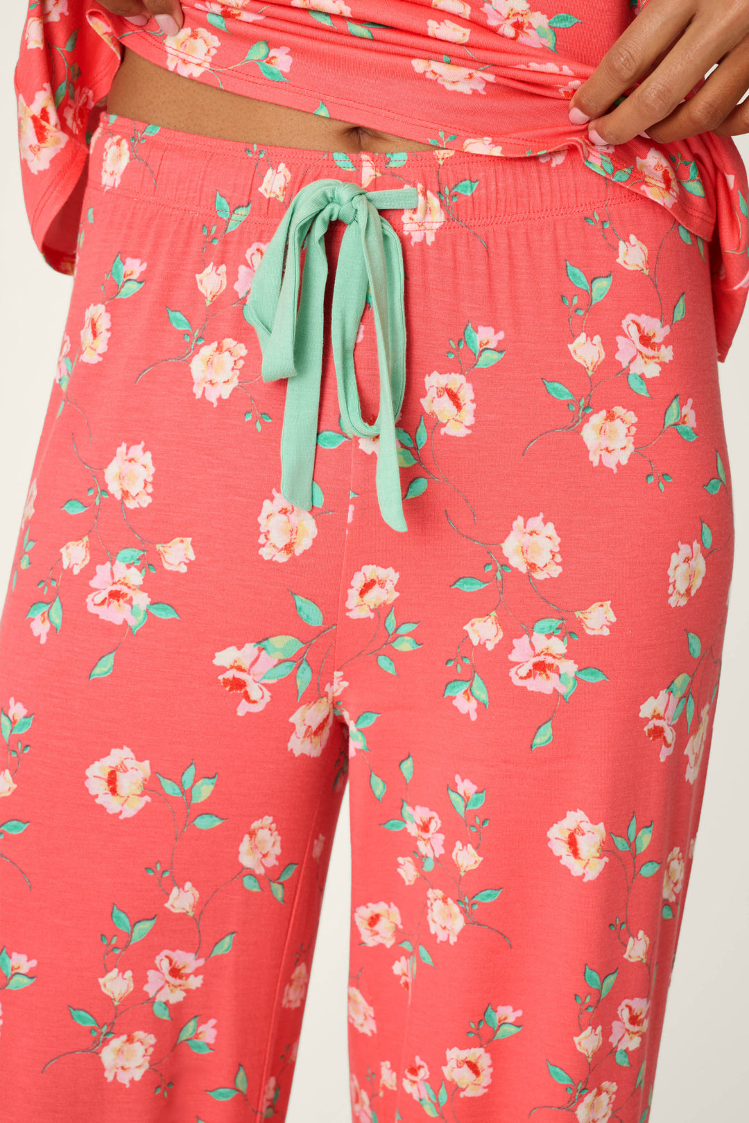 A person wears P.J. Salvage Siesta Dreams Dark Coral Floral Print Pajama Pants, featuring a wide-leg fit in modal jersey with a green drawstring and floral print. The matching top is slightly lifted to reveal the waistband.