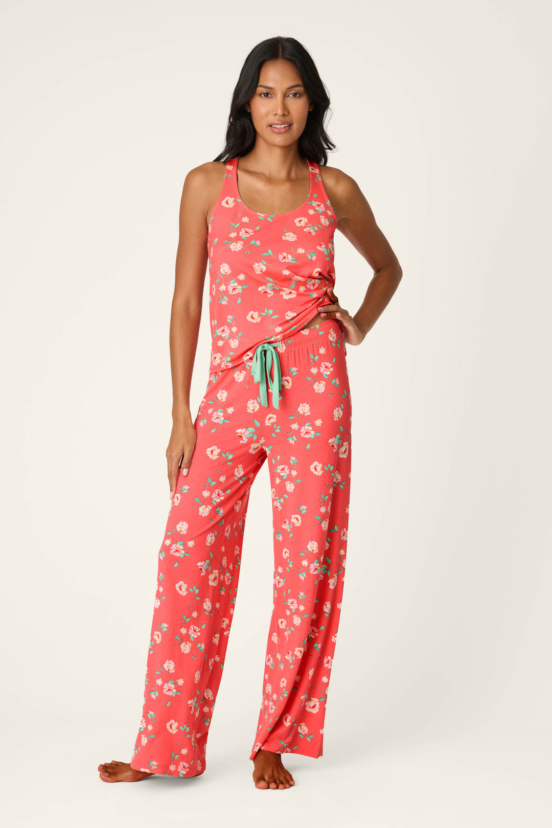A woman stands barefoot in P.J. Salvage's Siesta Dreams Dark Coral Floral Print Pajama Pants, crafted from soft modal jersey with white florals and a comfy drawstring waist, paired with a matching sleeveless tank top.
