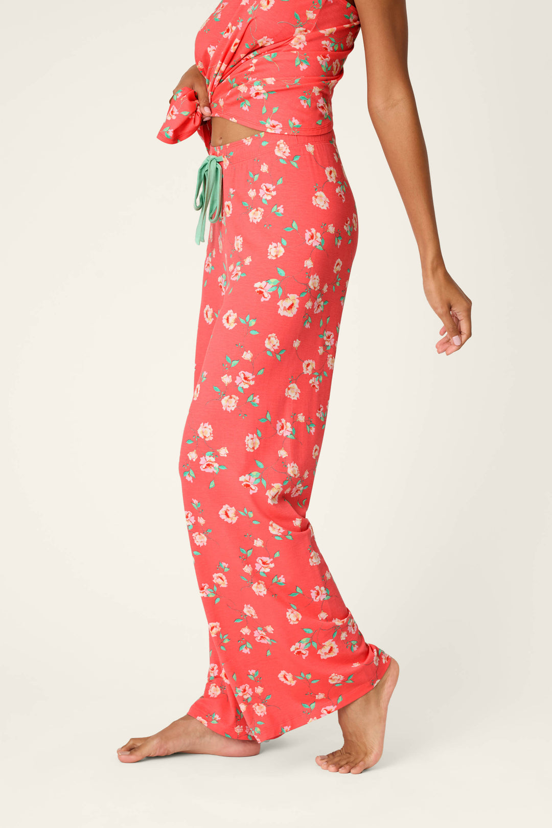 A person models P.J. Salvage's Siesta Dreams Dark Coral Floral Print Pajama Pants, standing barefoot on tiptoe and showing the side profile of the soft modal jersey pants against a light background.