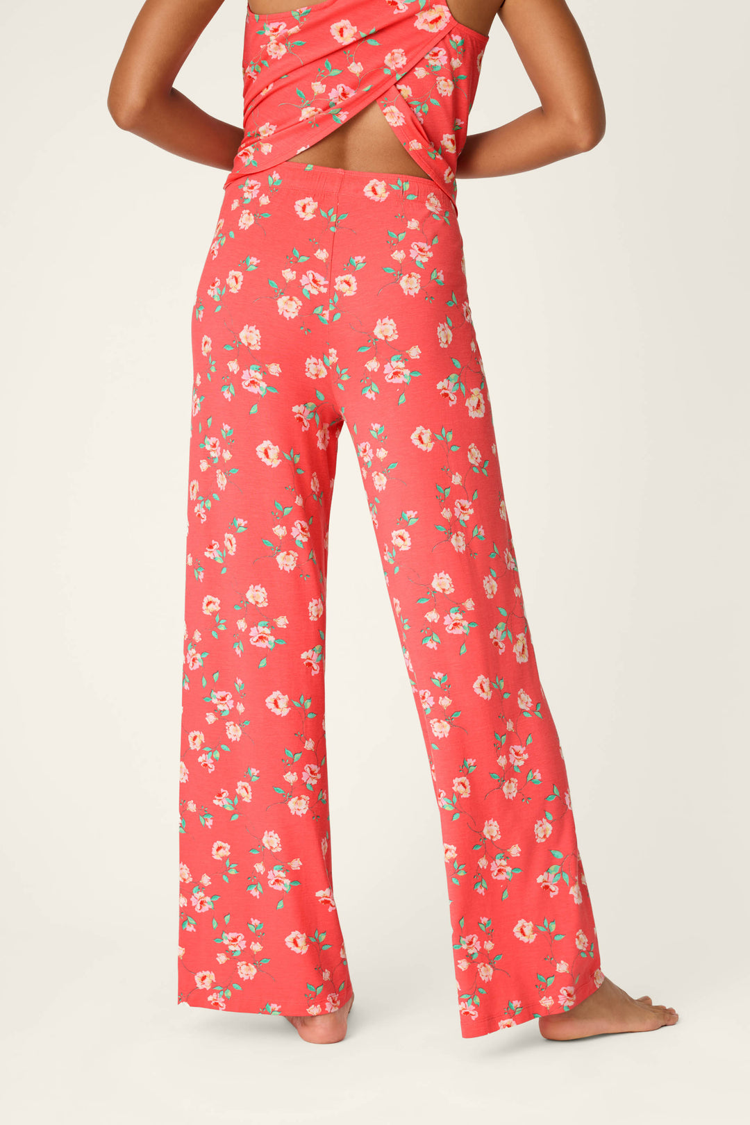 A person stands barefoot, facing away, wearing P.J. Salvage's Siesta Dreams Dark Coral Floral Print Pajama Pants and a matching backless crisscross top. The soft modal jersey drapes gently against a plain, light background.