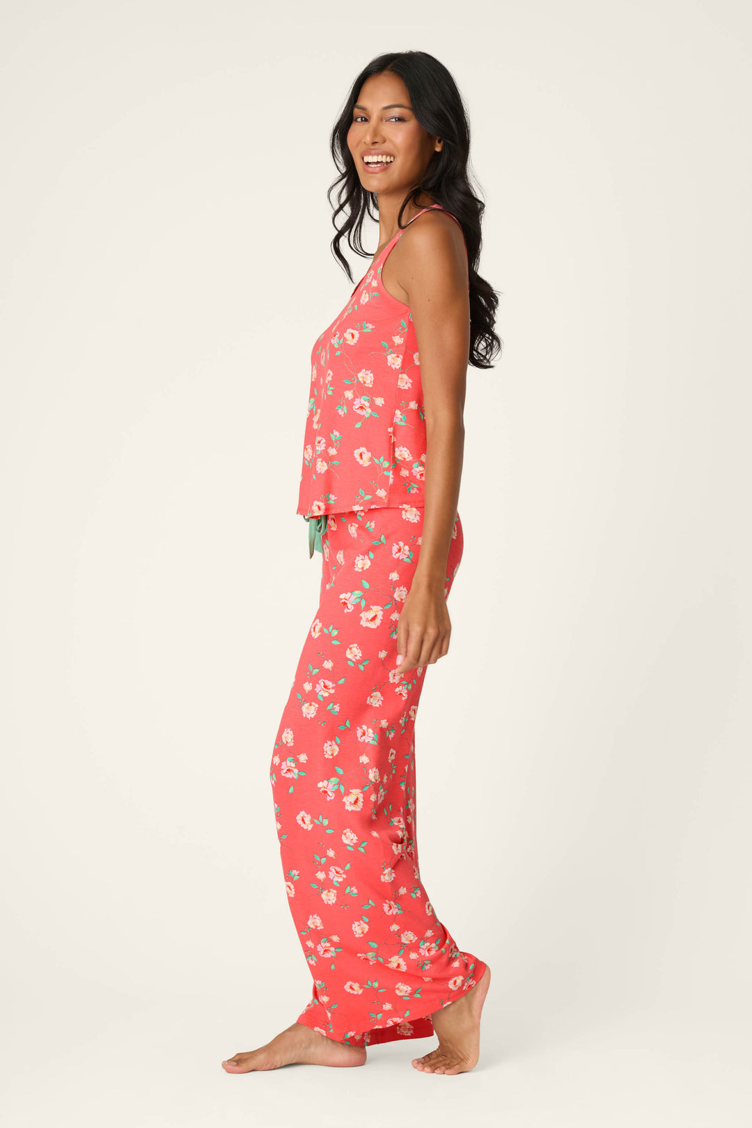 A woman smiles barefoot in the P.J. Salvage Siesta Dreams Dark Coral Floral Print Long PJ Set, featuring a sleeveless top and loose pants, as she poses against a plain light background.