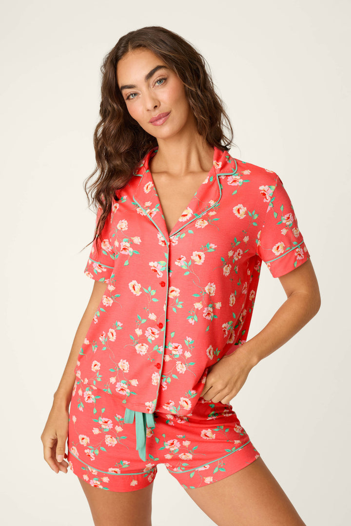 A woman with long, wavy brown hair wears the P.J. Salvage Siesta Dreams Dark Coral Floral Print Short Sleeve Top and matching shorts, standing with one hand on her hip against a plain light background.