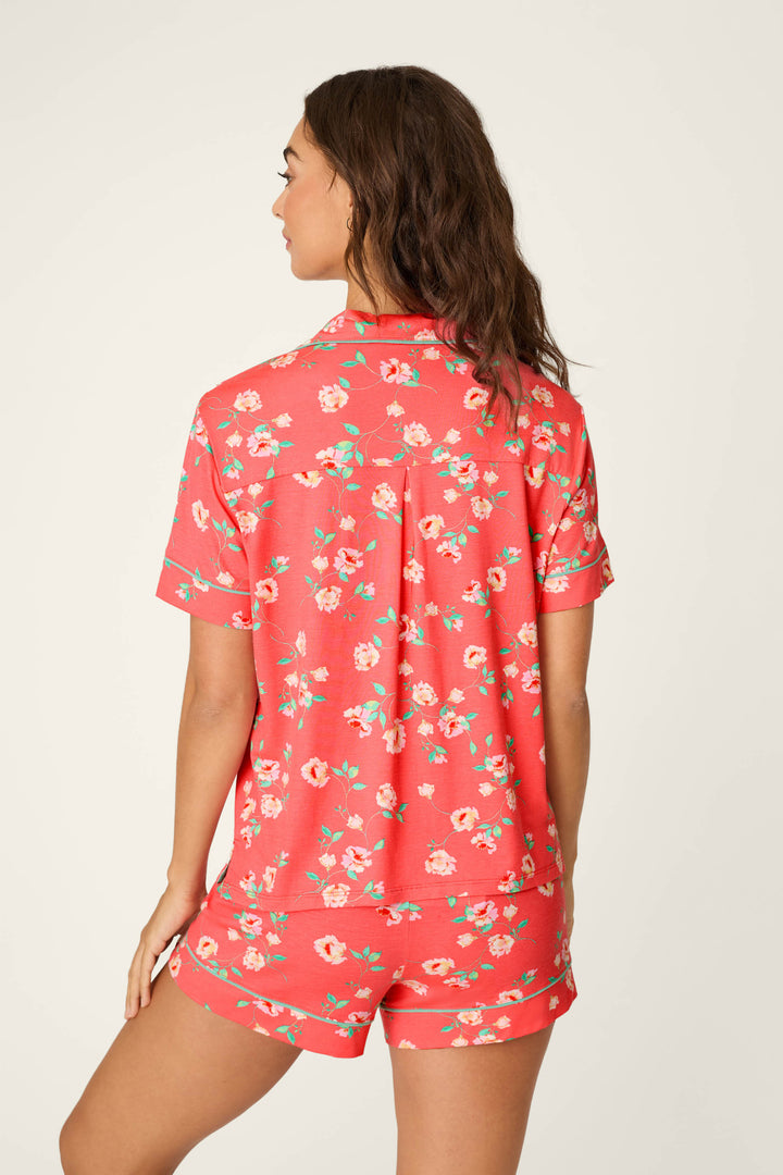 A woman with wavy brown hair faces away, wearing the P.J. Salvage Siesta Dreams Dark Coral Floral Print Short Sleeve Top and matching shorts. The modal jersey pajama set features a colorful floral print on a plain, light background.