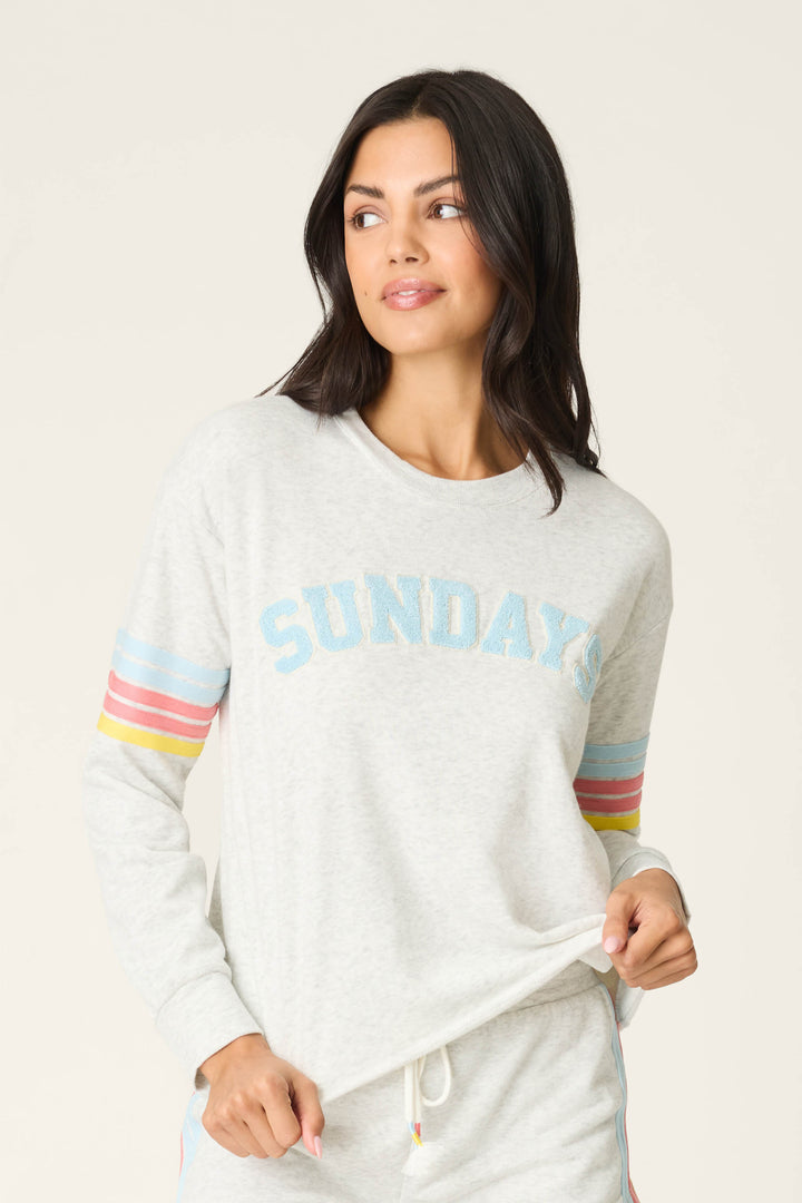 A woman with long dark hair wears the P.J. Salvage Sunday Funday Boucle "Sundays" Sweatshirt, a relaxed fit light gray fleece crew with blue lettering and rainbow sleeve stripes, looking to the side and holding the hem.