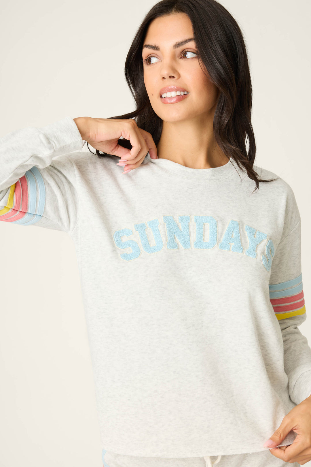 A woman wearing the P.J. Salvage Sunday Funday Boucle "Sundays" Sweatshirt, featuring blue lettering and pastel rainbow sleeve stripes, looks to the side and smiles with one hand touching her collar.