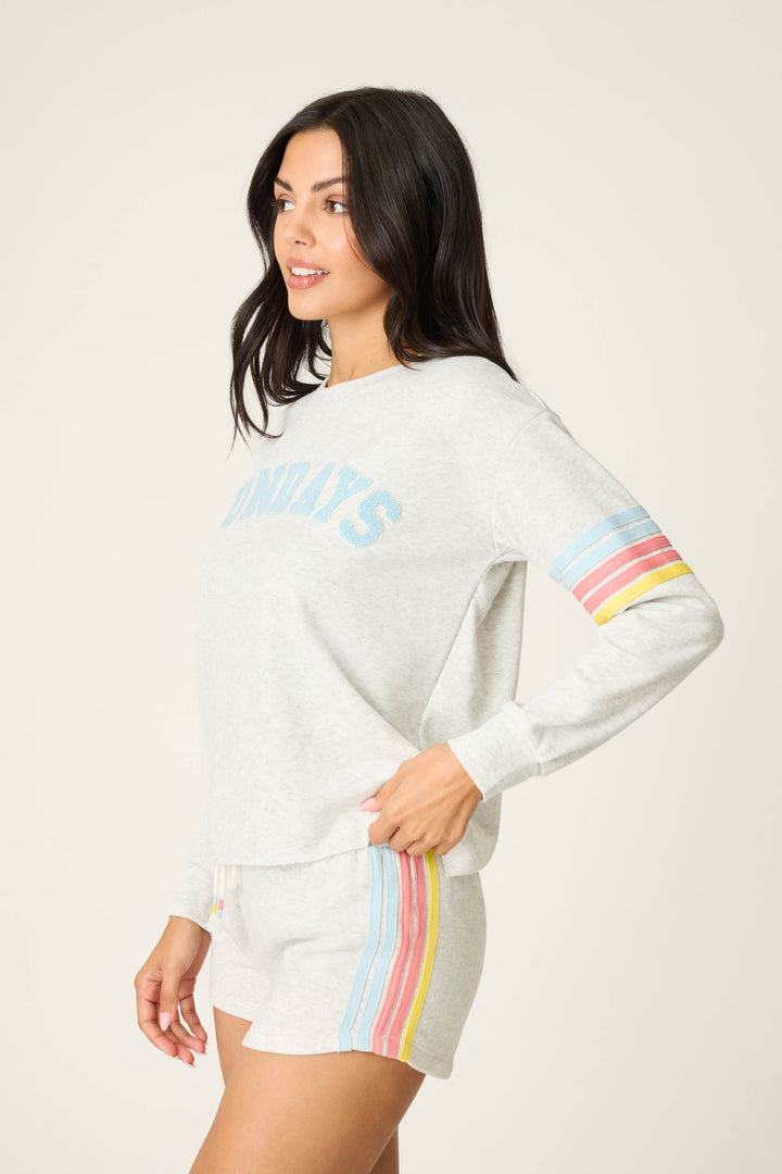 A woman with long dark hair wears the P.J. Salvage Sunday Funday Boucle "Sundays" Sweatshirt—a relaxed fit fleece in light gray with pastel rainbow stripes, blue "SUNDAYS" text, and matching shorts featuring colorful side details.