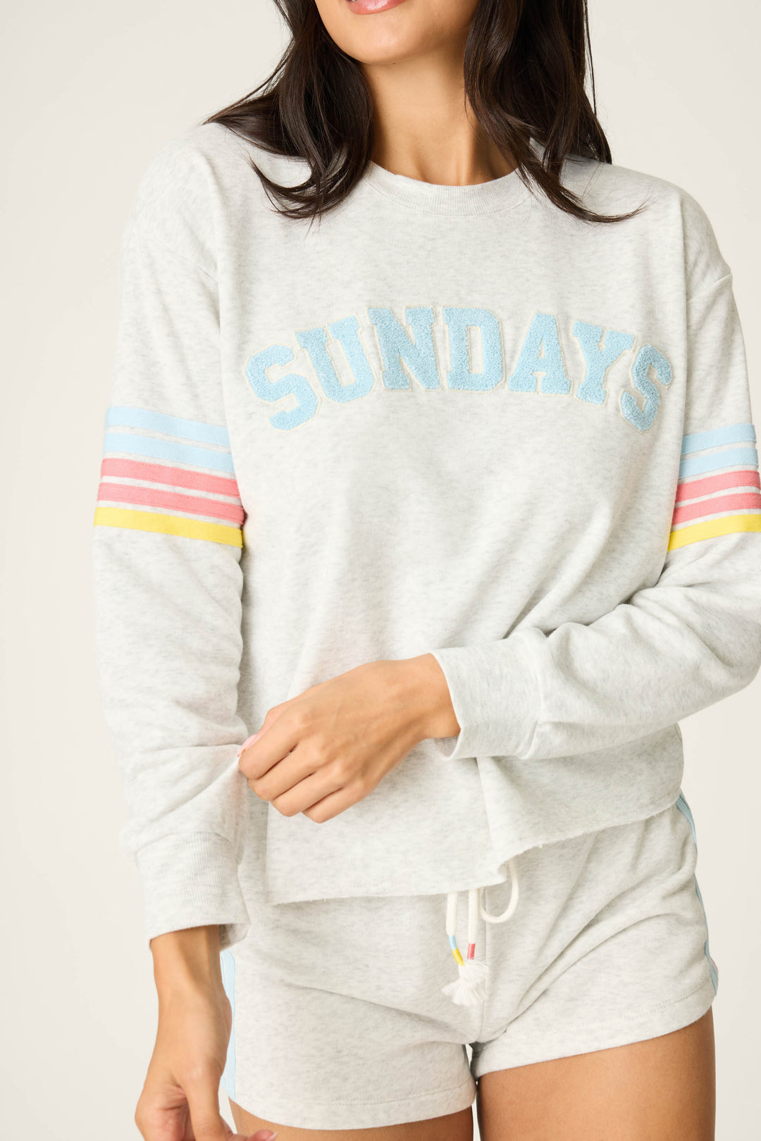 A woman wears the P.J. Salvage Sunday Funday Boucle "Sundays" Sweatshirt in relaxed fit, light gray, with blue lettering and pastel striped sleeves, paired with matching drawstring shorts. Her face is partially out of frame.