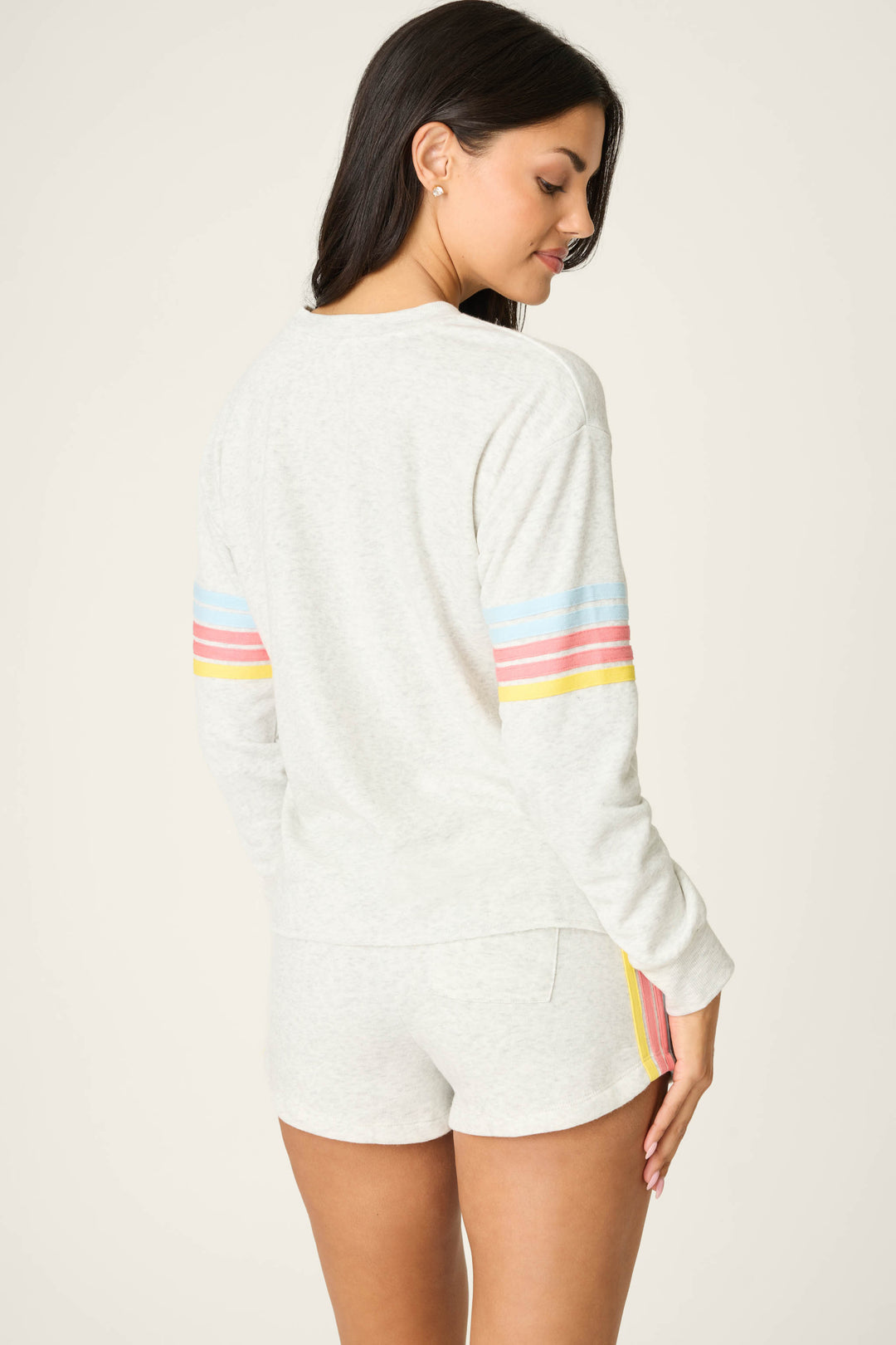 A woman with long dark hair stands with her back to the camera, wearing the P.J. Salvage Sunday Funday Boucle "Sundays" Sweatshirt in light gray with pastel rainbow striped sleeves and matching shorts, glancing over her shoulder and smiling slightly.