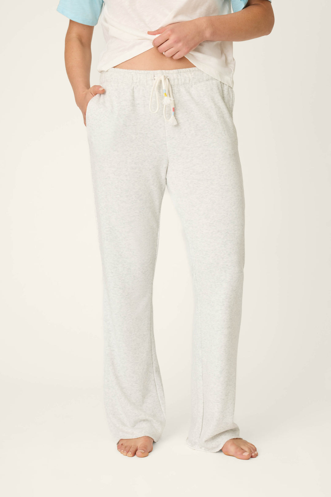 A person stands barefoot on a plain background, wearing P.J. Salvage Sunday Funday Open Leg Lounge Pants in light gray and a short-sleeve shirt, with one hand in the pocket and the shirt slightly lifted to show the waistband.