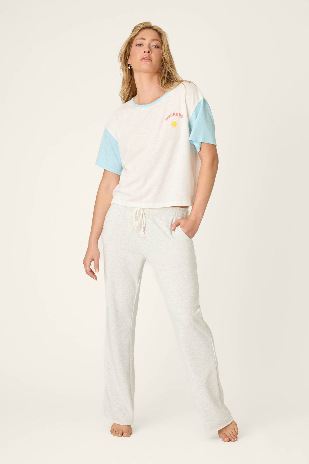A person stands barefoot in P.J. Salvage Sunday Funday Open Leg Lounge Pants and a white T-shirt with short light blue sleeves, featuring a bouclé sun detail and "WEEKEND" on the left chest, against a plain off-white background.