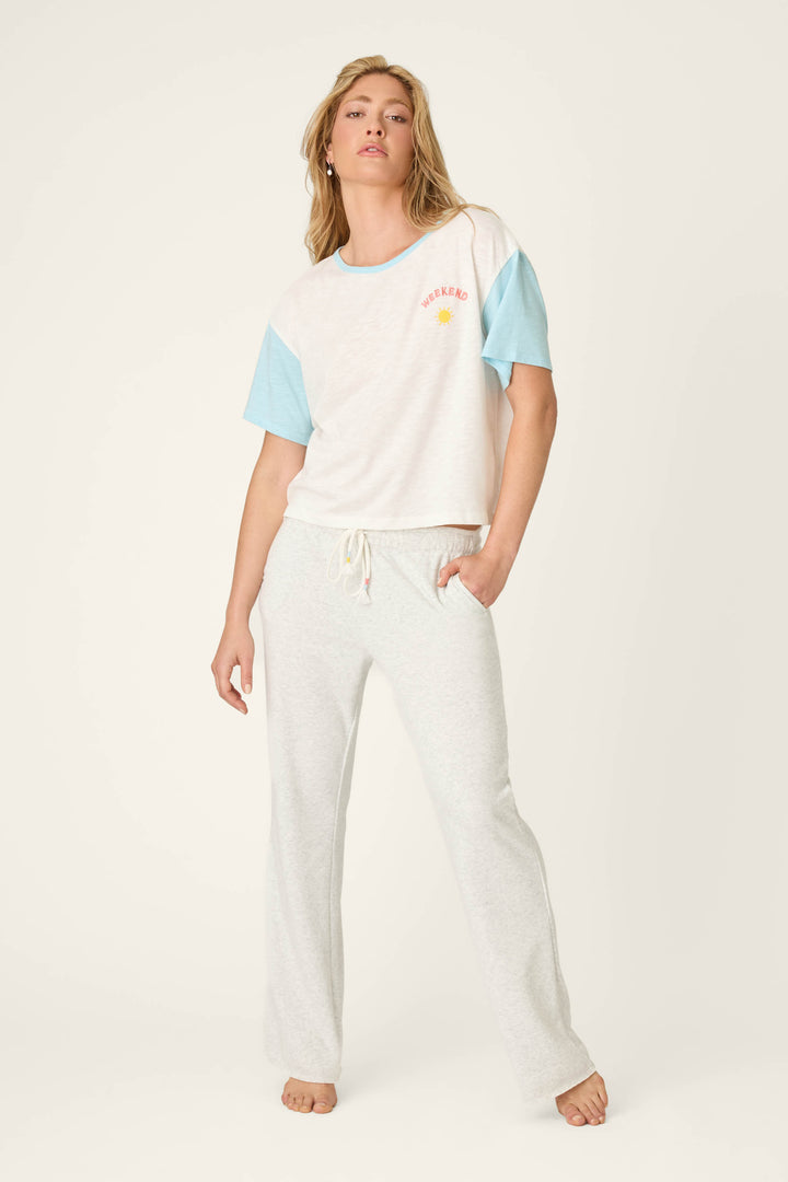 A person stands barefoot in P.J. Salvage Sunday Funday Open Leg Lounge Pants and a white T-shirt with short light blue sleeves, featuring a bouclé sun detail and "WEEKEND" on the left chest, against a plain off-white background.