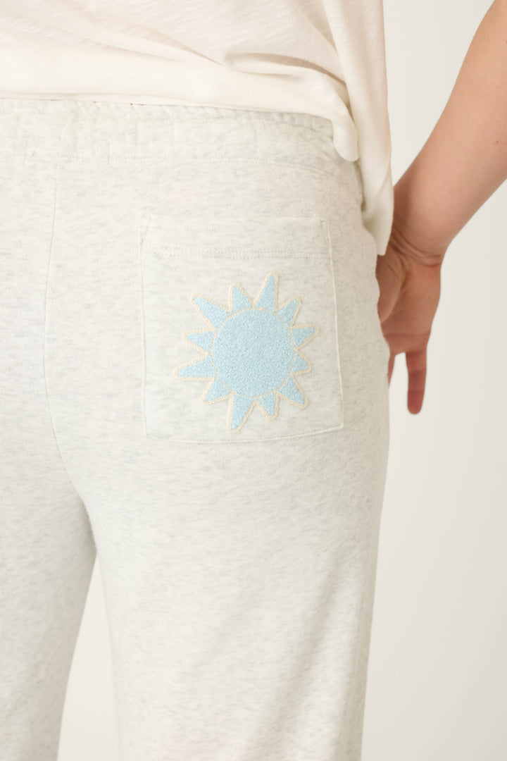 A person models the P.J. Salvage Sunday Funday Open Leg Lounge Pants in light gray, featuring a light blue and yellow sun embroidered on the back pocket. Only their lower back and upper legs are shown.