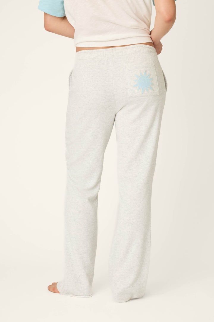 A person stands barefoot, facing away, wearing P.J. Salvage’s Sunday Funday Open Leg Lounge Pants in light gray with a bouclé sun detail on the back pocket and a short-sleeve shirt with pale blue sleeves against an off-white background.