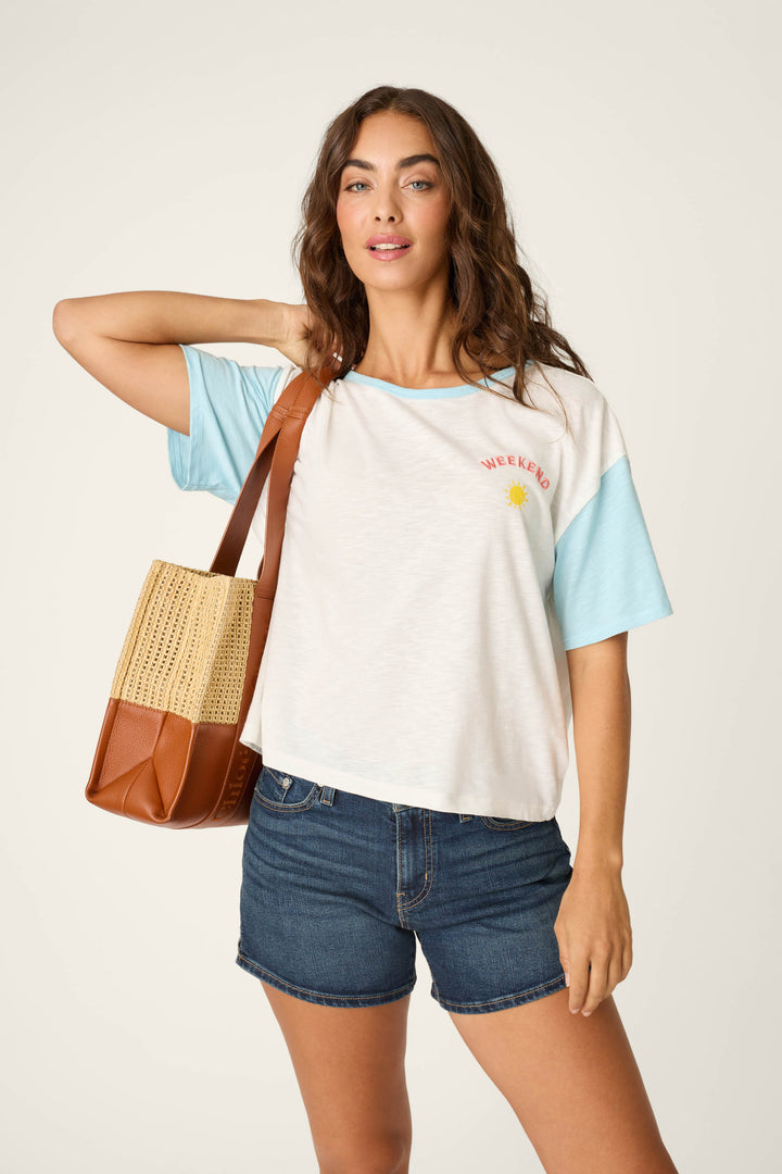 A woman poses against a light background, wearing P.J. Salvage's Sunday Funday Embroidered "Weekend" T-Shirt in white and light blue with a sun graphic, denim shorts, and carrying a brown woven tote over her shoulder.