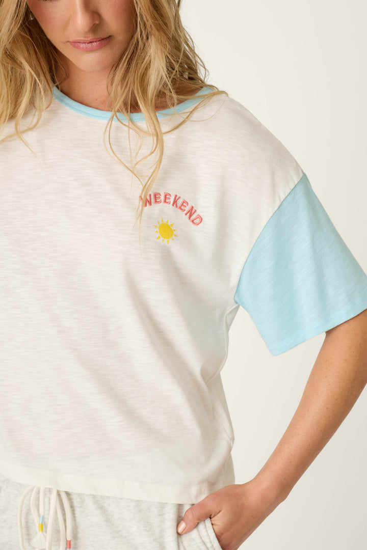 A woman wears the P.J. Salvage Sunday Funday Embroidered "Weekend" T-Shirt—a white slub jersey with light blue sleeves and a sun graphic—standing with one hand in her light-colored pants pocket, upper body visible.