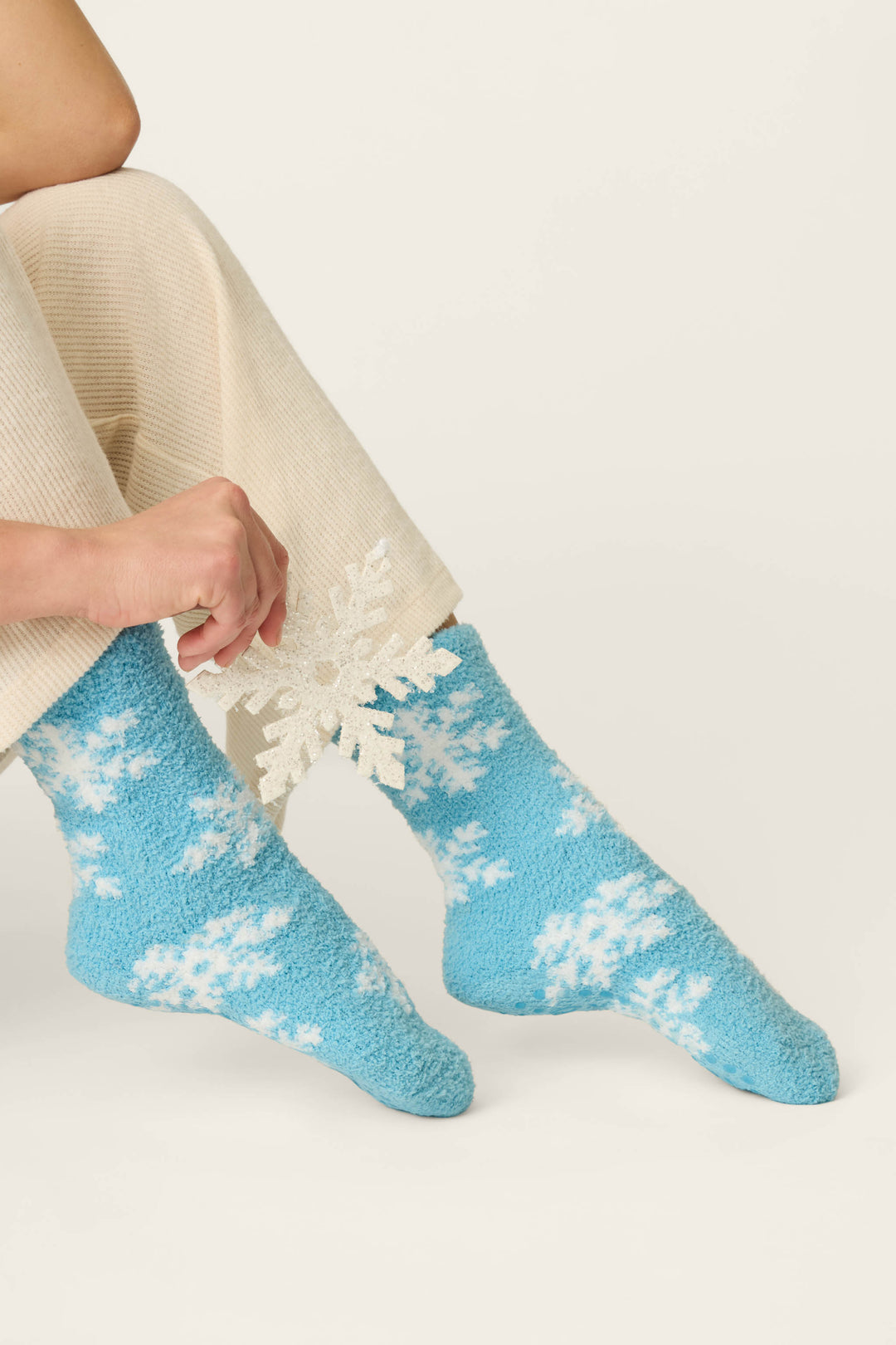 A person wearing cream pants and P.J. Salvage Ski Jammie Classics Snowflake Print Socks sits while holding a white paper snowflake.