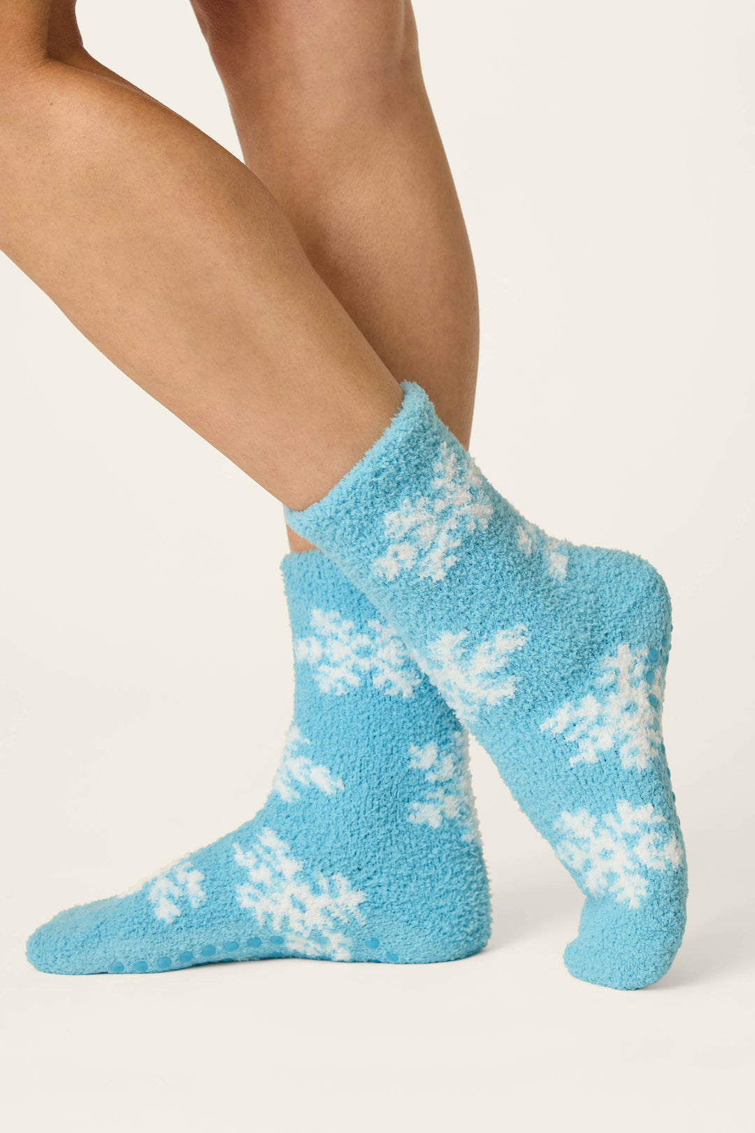 A pair of legs wearing P.J. Salvage Ski Jammie Classics Snowflake Print Socks, featuring a fluffy blue design with white snowflake patterns, set against a plain light background.