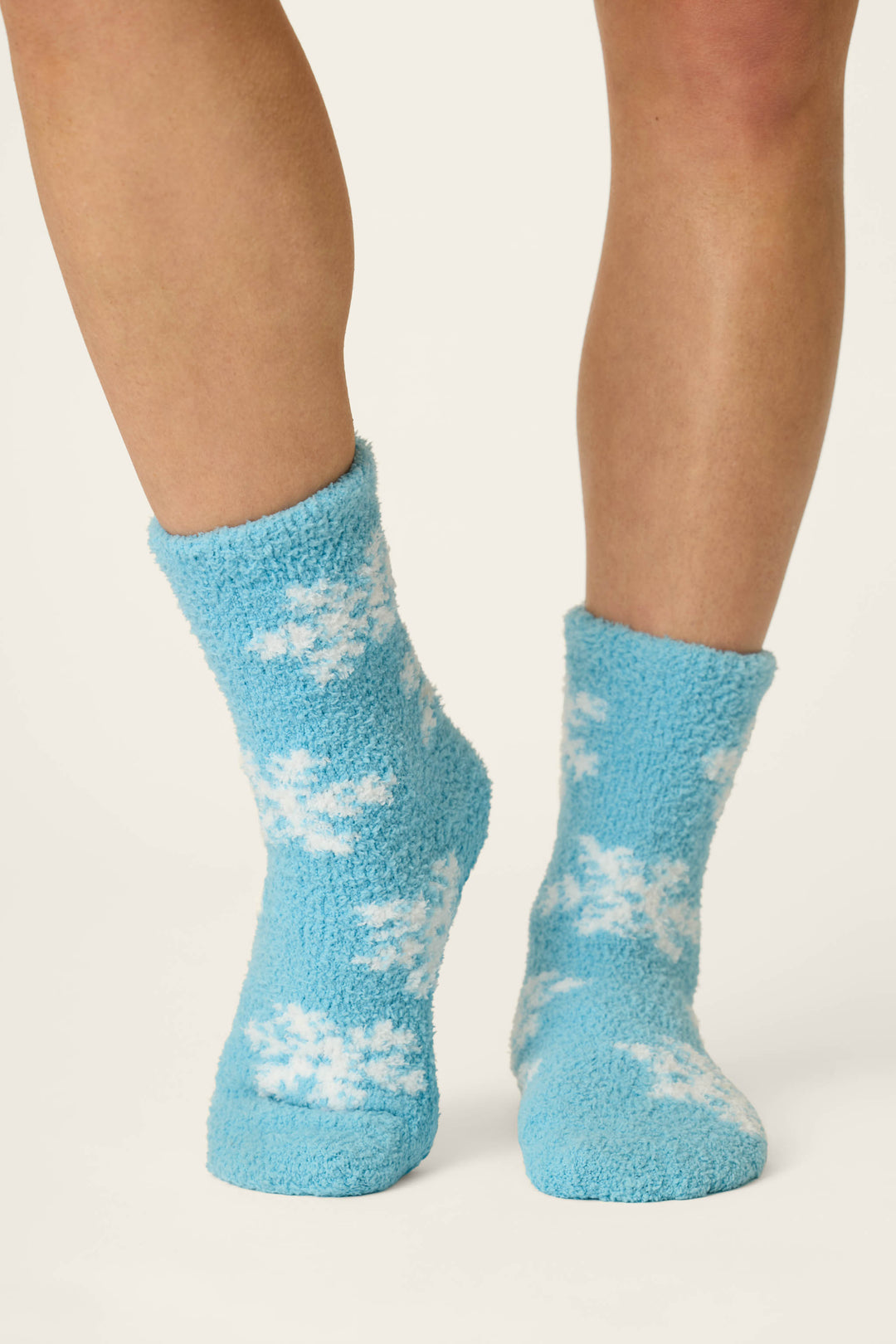 A person wearing P.J. Salvage Ski Jammie Classics Snowflake Print Socks, featuring a fuzzy blue design with white snowflakes, stands on a light background. Only the lower legs and feet are visible.