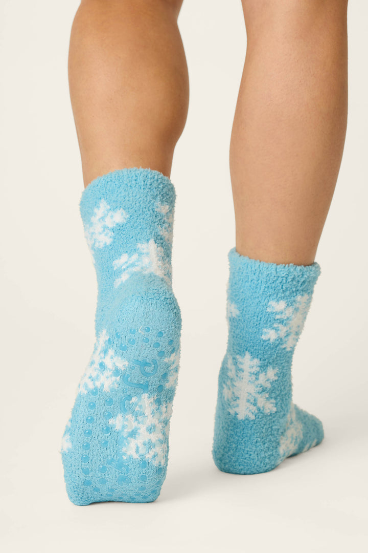 A person is shown from the back wearing P.J. Salvage Ski Jammie Classics Snowflake Print Socks—fuzzy blue socks featuring white snowflake designs. The soft, cozy socks cover the ankles and part of the lower legs, set against a plain white background.