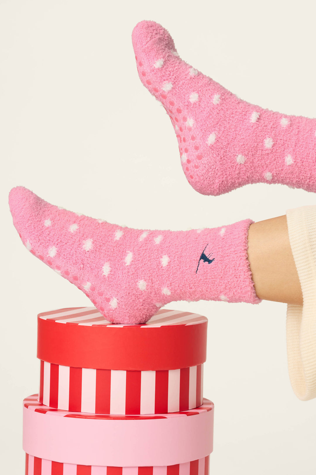 Two feet wearing P.J. Salvage's Ski Jammie Classics Ski Bunny & Snowflakes Socks rest on red and white striped round boxes. The background is plain and light-colored.