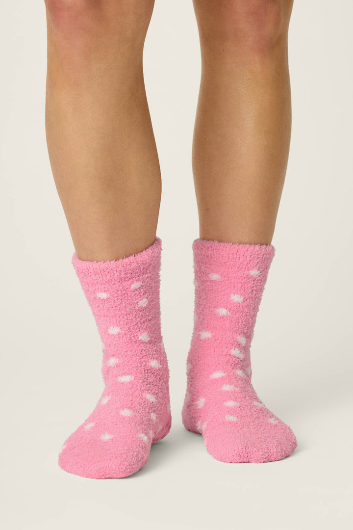 A pair of legs wearing P.J. Salvage Ski Jammie Classics Ski Bunny & Snowflakes Socks, featuring a fuzzy pink design with white polka dots, standing on a light-colored surface.