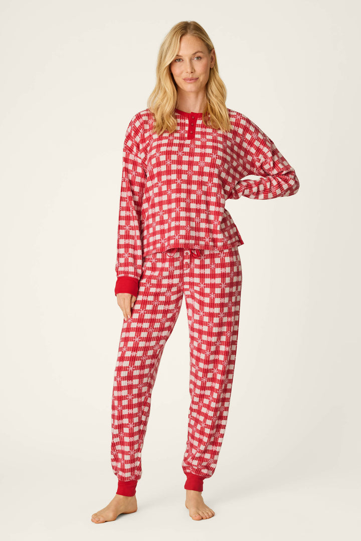 A woman with blonde hair stands barefoot against a plain light background, smiling softly as she wears the Ski Jammie Classics Snowflake Picnic Jogger PJ Set by P.J. Salvage—a red and white checkered pajama set with red cuffs.