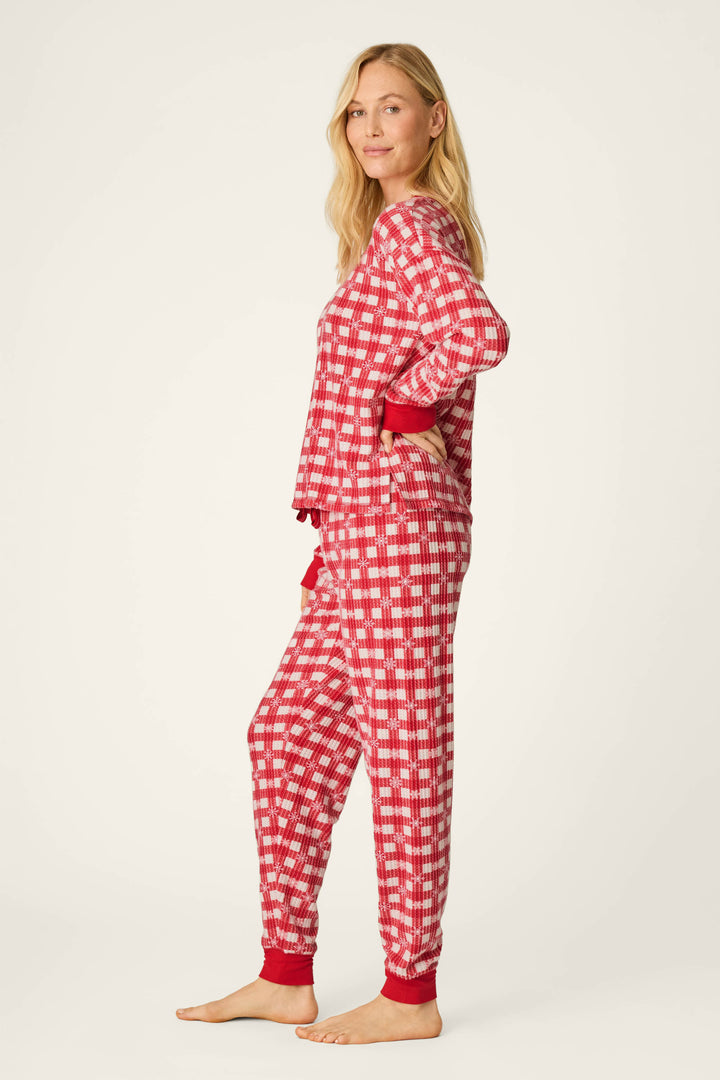 A woman with long blonde hair stands barefoot in side profile, wearing the P.J. Salvage Ski Jammie Classics Snowflake Picnic Jogger PJ Set—red and white checkered pajamas with red cuffs—posed against a plain, light background.