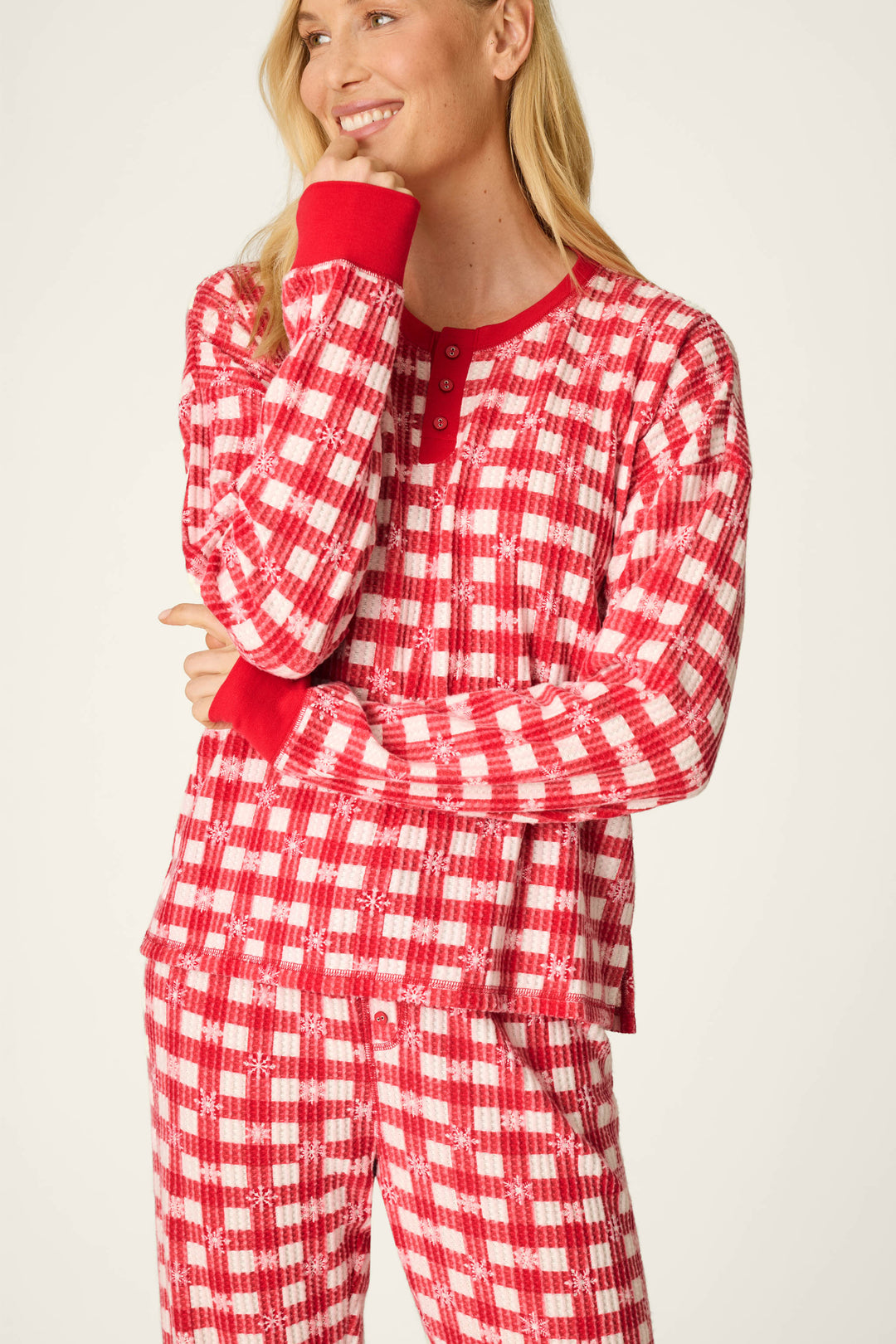 A smiling woman wears the P.J. Salvage Ski Jammie Classics Snowflake Picnic Jogger PJ Set, featuring red and white plaid with red cuffs, standing with one hand touching her face and the other arm crossed.