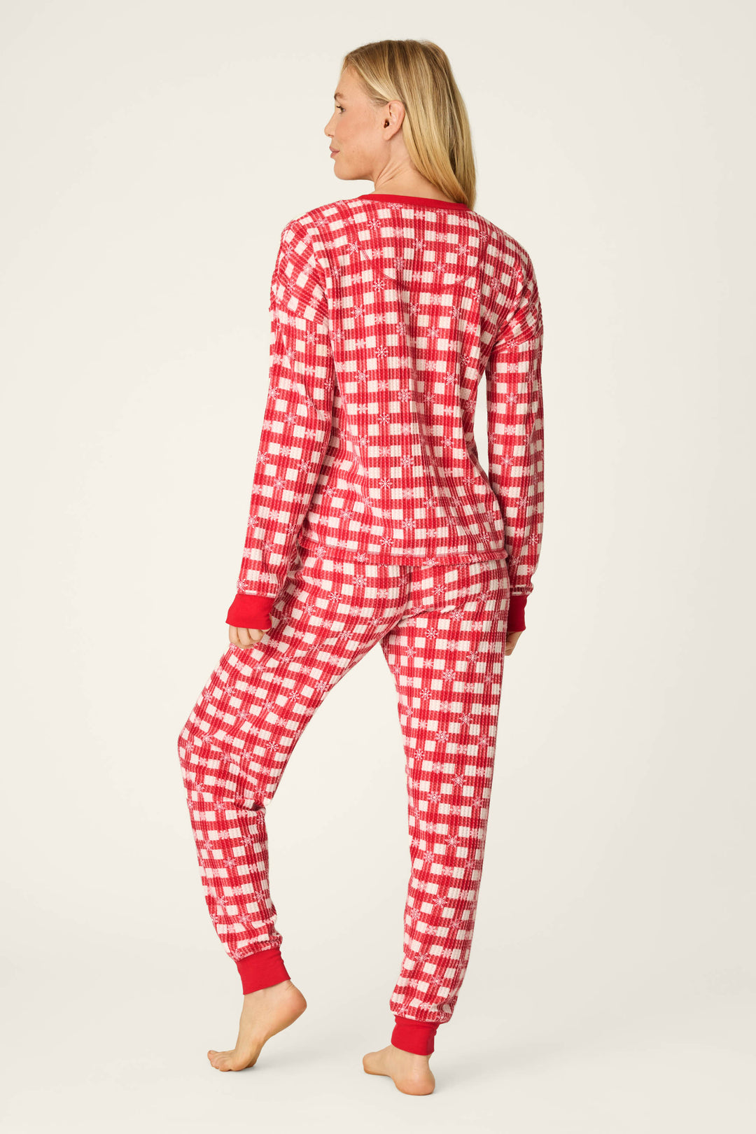 A person stands barefoot, facing away from the camera, wearing the P.J. Salvage Ski Jammie Classics Snowflake Picnic Jogger PJ Set— a red and white plaid long-sleeve pajama set with red cuffs—against a plain light background.