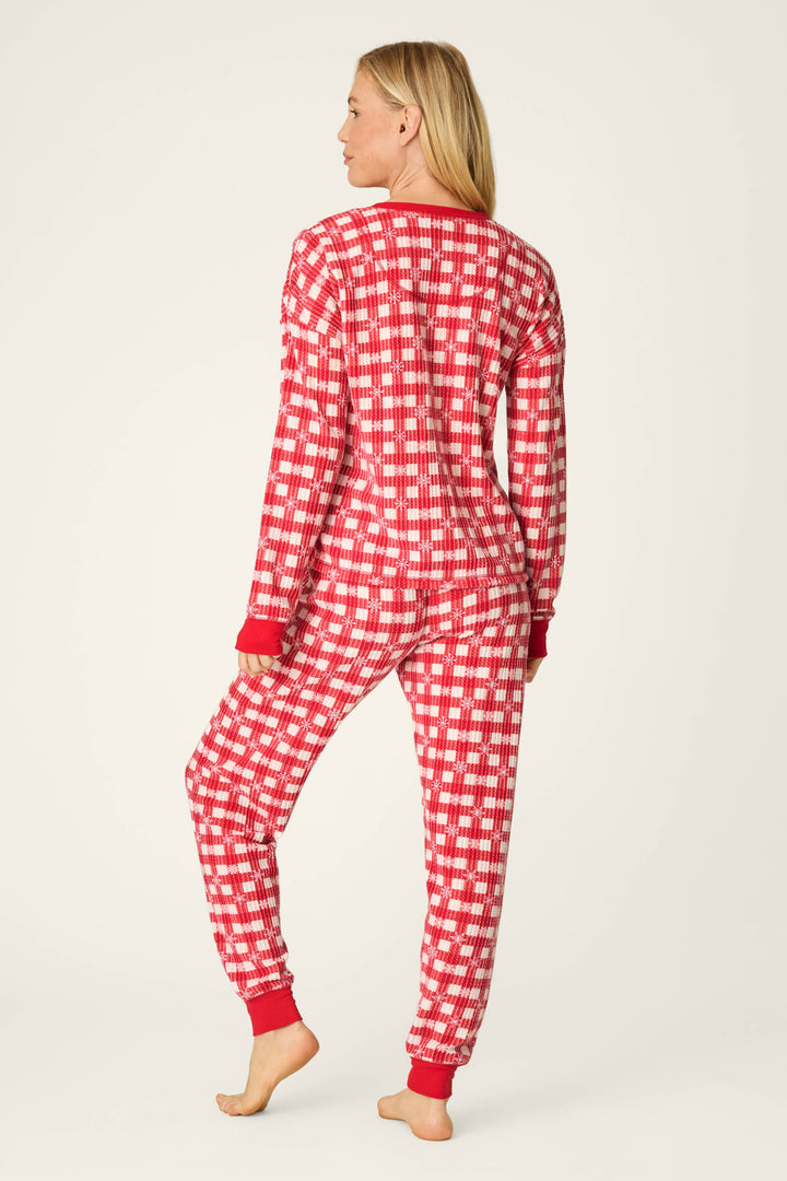 A person stands barefoot, facing away from the camera, wearing the P.J. Salvage Ski Jammie Classics Snowflake Picnic Jogger PJ Set— a red and white plaid long-sleeve pajama set with red cuffs—against a plain light background.