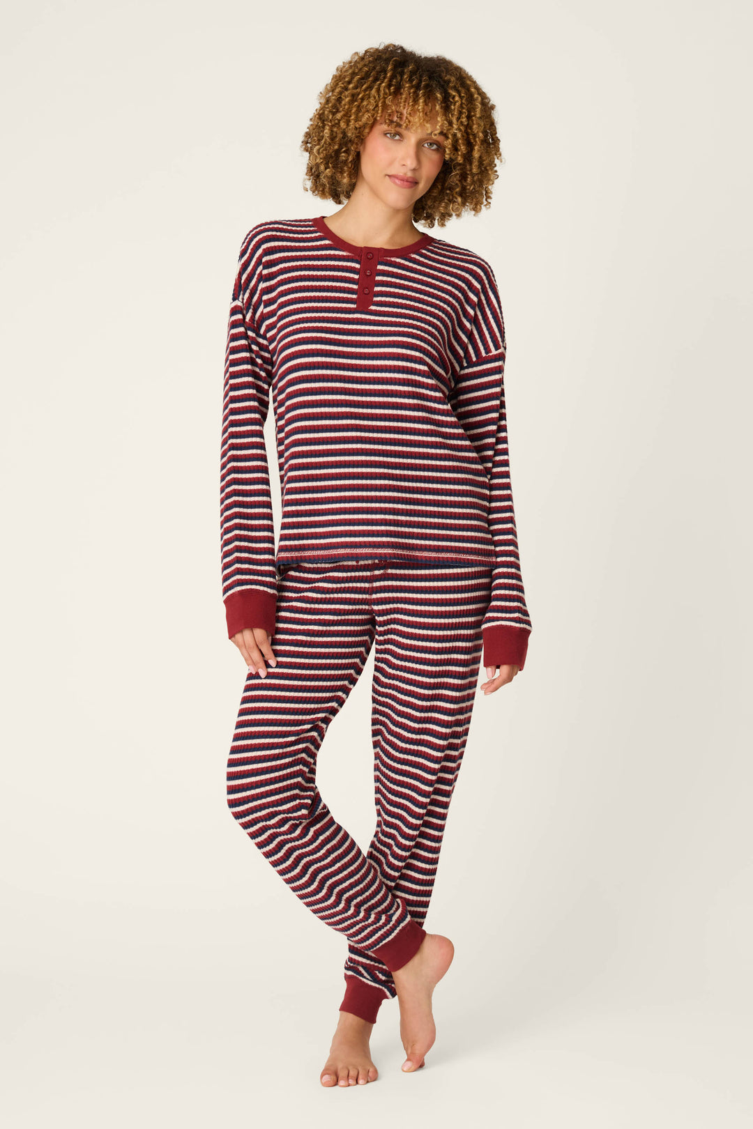 A person models the P.J. Salvage Ski Jammie Classics Striped Long Sleeve Jogger PJ Set, featuring maroon and white stripes, as they stand barefoot on a white background with one leg crossed in front of the other and a relaxed expression.
