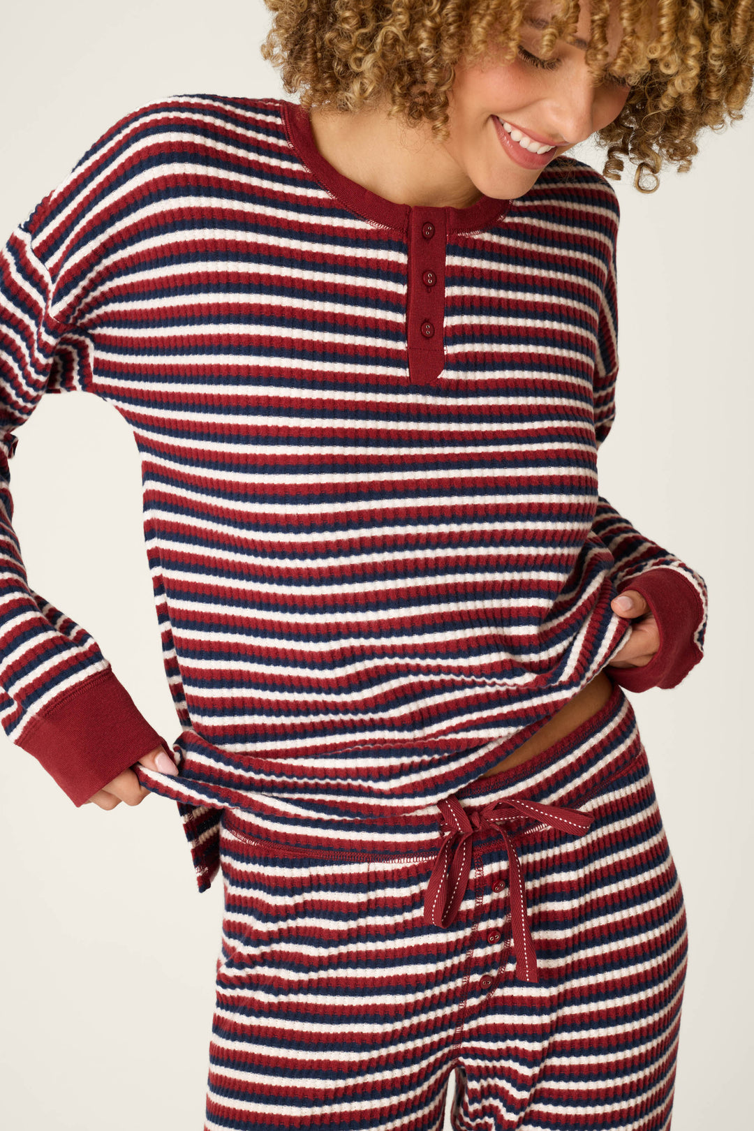 A person wearing the P.J. Salvage Ski Jammie Classics Striped Long Sleeve Jogger PJ Set smiles and lifts the shirt slightly to reveal the waistband of the matching pants, showcasing the cozy red, white, and navy stripes.