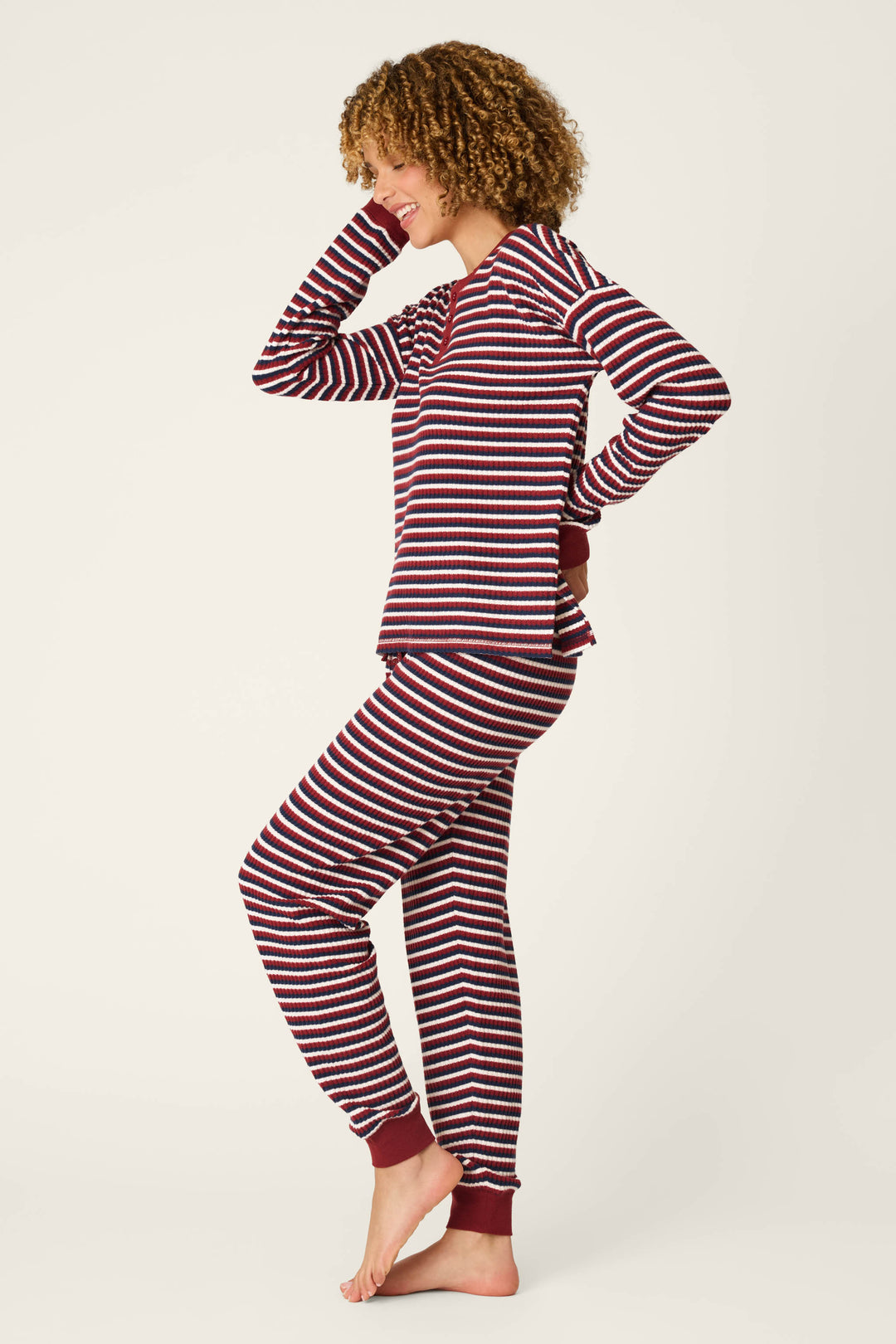 Wearing the P.J. Salvage Ski Jammie Classics Striped Long Sleeve Jogger PJ Set, a person stands barefoot against a plain background, smiling with one hand on their hip and the other touching their head.
