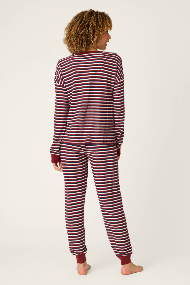 A person stands barefoot, facing away, in front of a plain light background, wearing the P.J. Salvage Ski Jammie Classics Striped Long Sleeve Jogger PJ Set—a matching top and pants featuring horizontal maroon and white stripes with maroon cuffs.