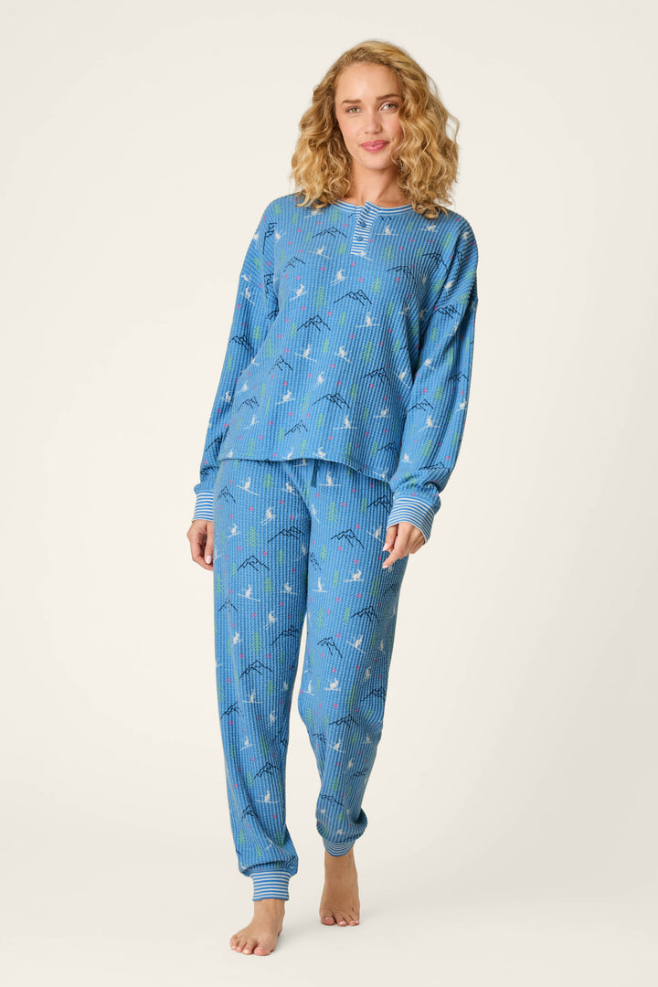 A woman with curly blonde hair wears the P.J. Salvage Ski Jammie Classics Skiing Bunnies Long Sleeve Jogger PJ Set. She stands barefoot on a plain background, smiling gently with one knee slightly bent and her hands relaxed at her sides.