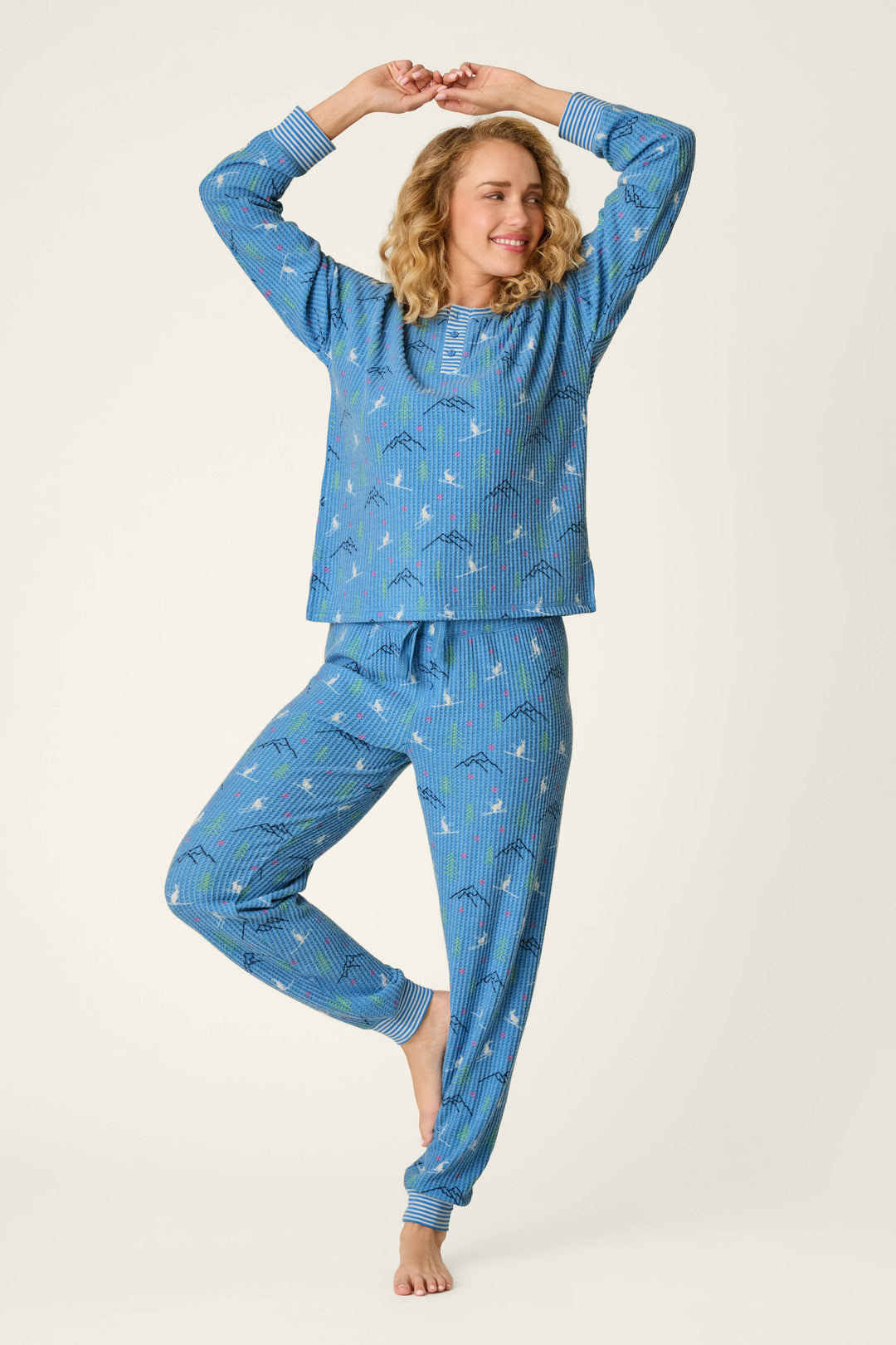 A smiling person with curly hair wears the P.J. Salvage Ski Jammie Classics Skiing Bunnies Long Sleeve Jogger PJ Set, standing on one leg with arms raised and happily posing against a plain light background.