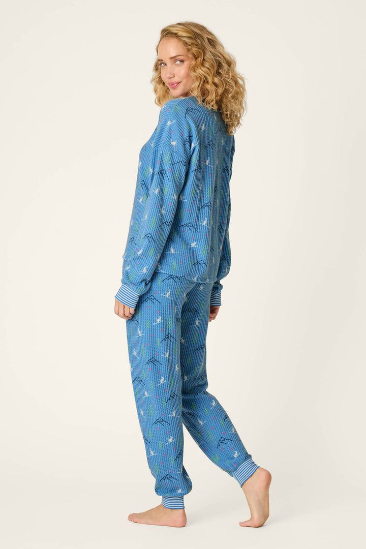 A person with curly blonde hair wears the Ski Jammie Classics Skiing Bunnies Long Sleeve Jogger PJ Set by P.J. Salvage, standing barefoot and turned slightly to the side, looking over their shoulder and smiling against a plain light background.