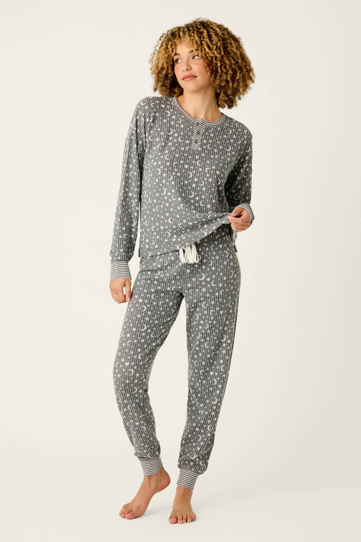 A person wearing the P.J. Salvage Ski Jammie Classics Celestial Print Long Sleeve Jogger PJ Set stands barefoot on a light background, gently holding the hem of the top away from their body.