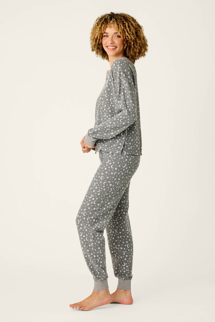 A smiling person with curly hair stands sideways, barefoot on a plain light background, wearing the P.J. Salvage Ski Jammie Classics Celestial Print Long Sleeve Jogger PJ Set—a matching gray pajama set featuring a white geometric pattern.