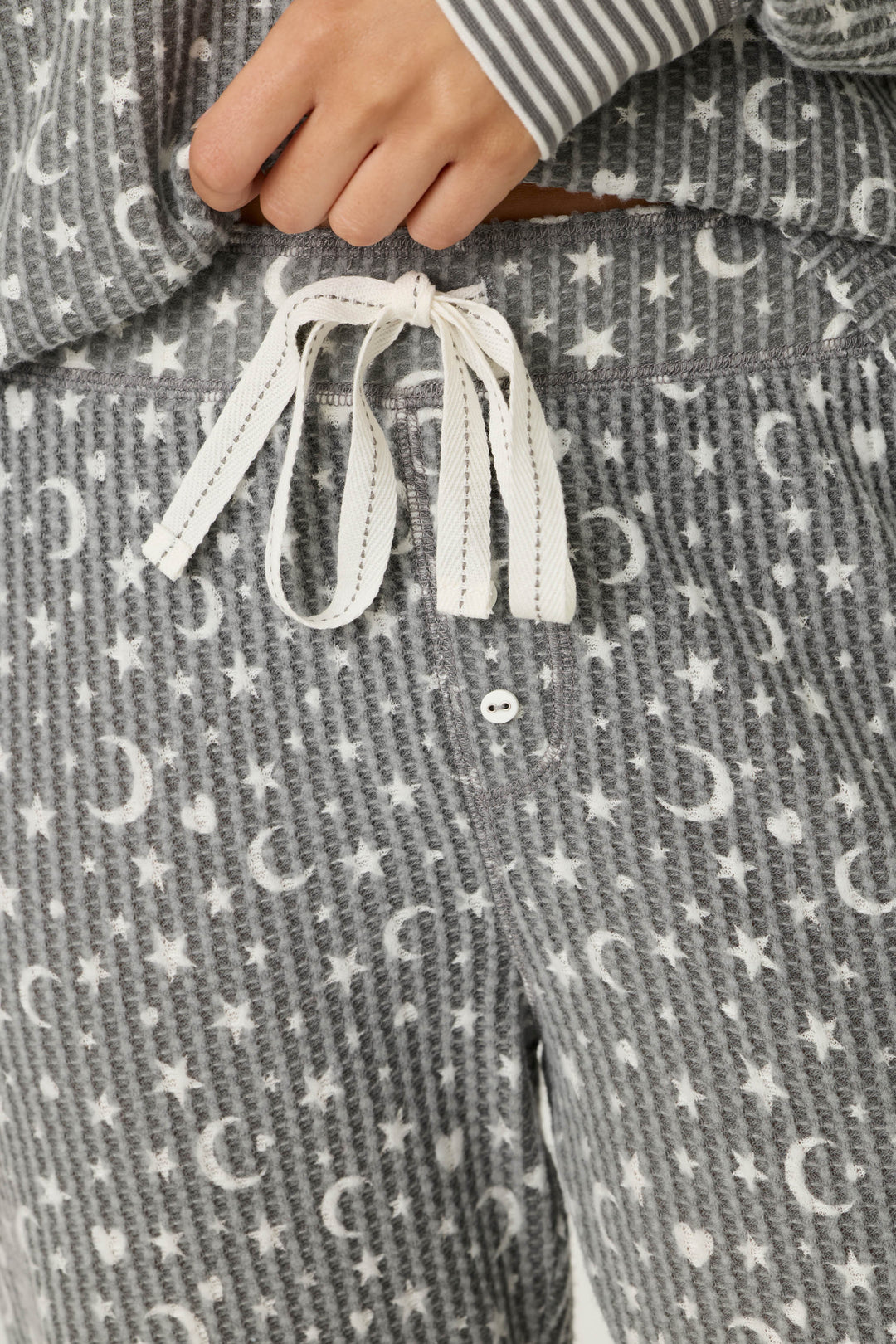 A close-up of a person wearing the P.J. Salvage Ski Jammie Classics Celestial Print Long Sleeve Jogger PJ Set, featuring grey pajama pants with white stars and moons, a white drawstring, and a matching top with striped cuffs.