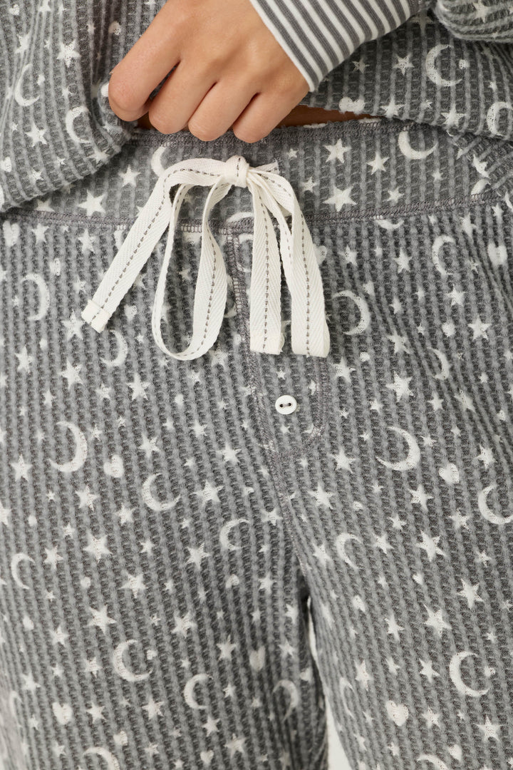 A close-up of a person wearing the P.J. Salvage Ski Jammie Classics Celestial Print Long Sleeve Jogger PJ Set, featuring grey pajama pants with white stars and moons, a white drawstring, and a matching top with striped cuffs.