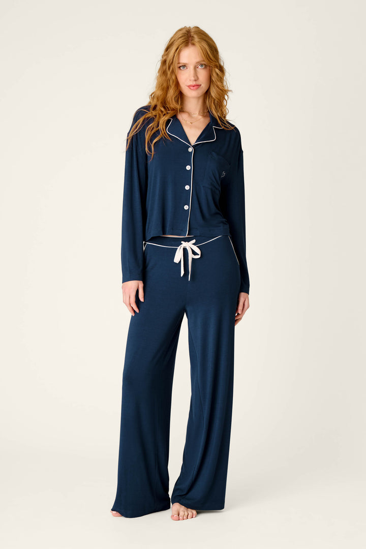 A woman with long red hair wears the Soiree Dreams Deep Navy Long Sleeve PJ Set by P.J. Salvage, featuring white trim and buttons, as she stands barefoot against a plain light background.
