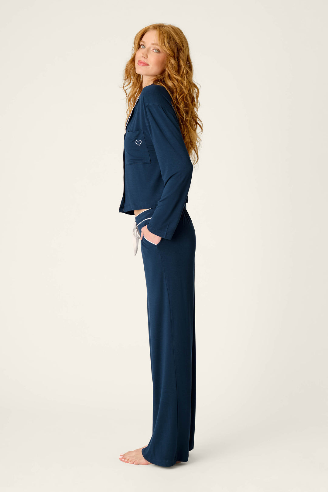 A person with long wavy hair stands barefoot in profile, wearing the P.J. Salvage Soiree Dreams Deep Navy Long Sleeve PJ Set with hands in pockets, against a plain light background.